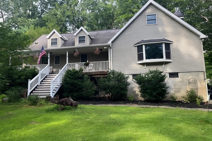 New england style home featuring a front lawn, a porch, stairs, and view of wooded area
