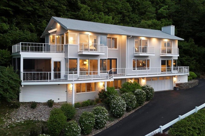Back of house with a balcony, a chimney, driveway, an attached garage, and a forest view