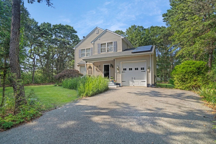 Traditional home featuring gravel driveway, roof mounted solar panels, a garage, a front lawn, and view of wooded area