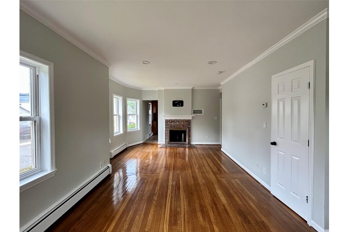 Unfurnished living room featuring baseboard heating, dark wood finished floors, a brick fireplace, and ornamental molding