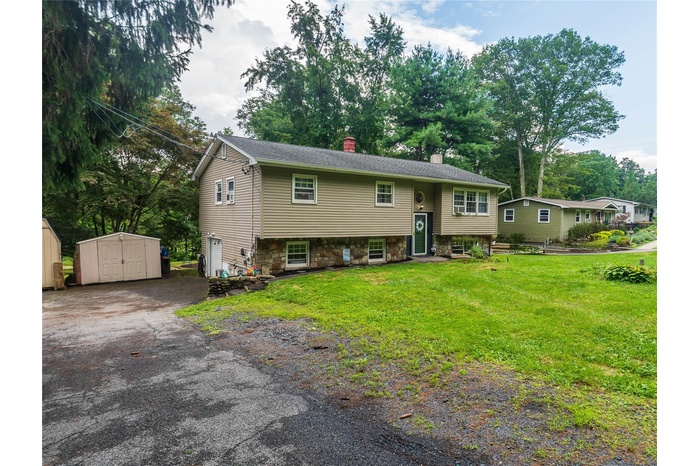 Raised ranch featuring a storage unit, a chimney, driveway, stone siding, and a front yard