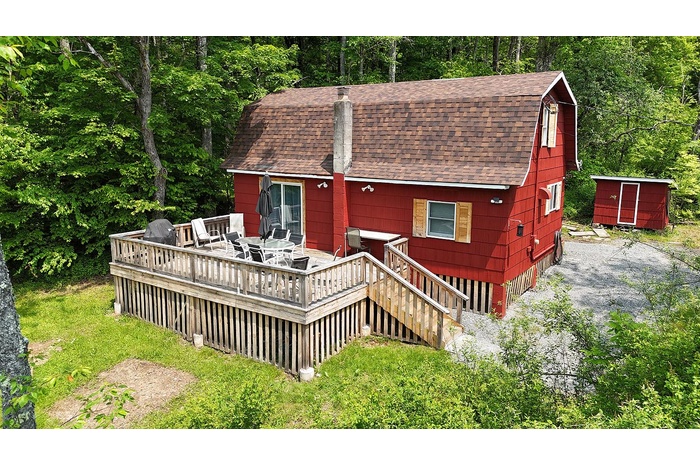 Rear view of property featuring a shingled roof, a deck, a gambrel roof, a view of trees, and an outdoor structure