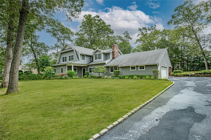 Shingle-style home featuring a chimney, a shingled roof, driveway, and a front lawn