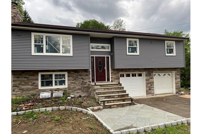 Raised ranch with driveway, a garage, stone siding, entry steps, and a chimney