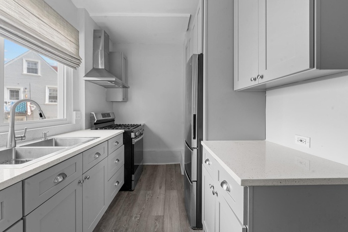 Kitchen featuring appliances with stainless steel finishes, wall chimney exhaust hood, gray cabinetry, wood finished floors, and light stone counters