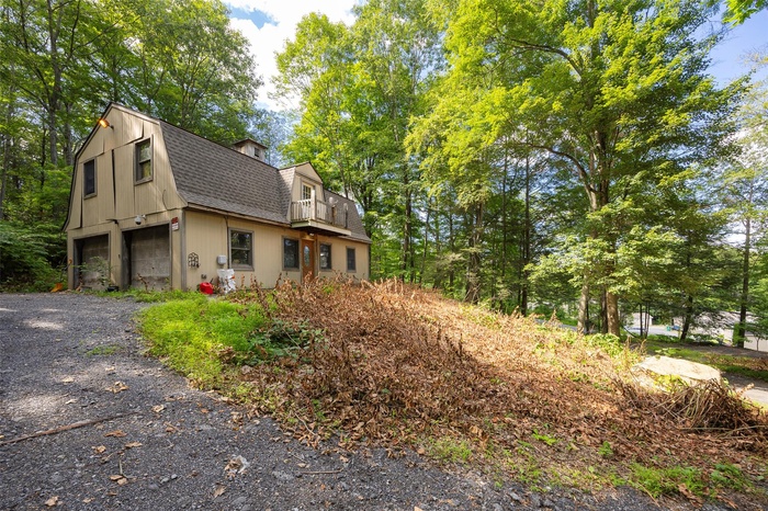 View of home's exterior featuring a gambrel roof, a balcony, roof with shingles, driveway, and view of scattered trees