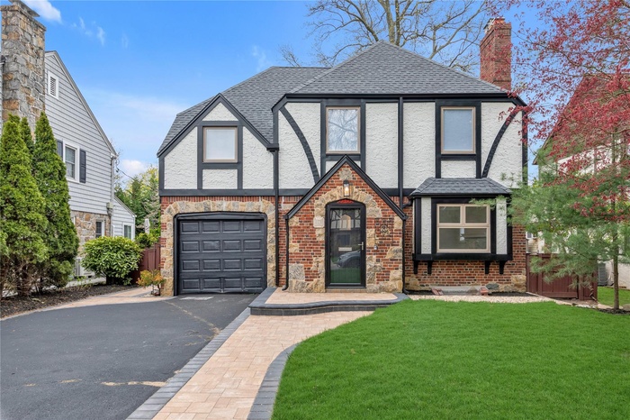 Tudor home with a front lawn, stucco siding, an attached garage, driveway, and roof with shingles