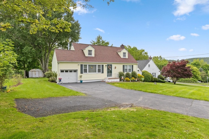 Cape cod home with driveway, a chimney, a front yard, a garage, and a shingled roof