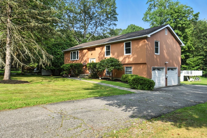 View of front facade with driveway, a front lawn, a shingled roof, an attached garage, and view of wooded area