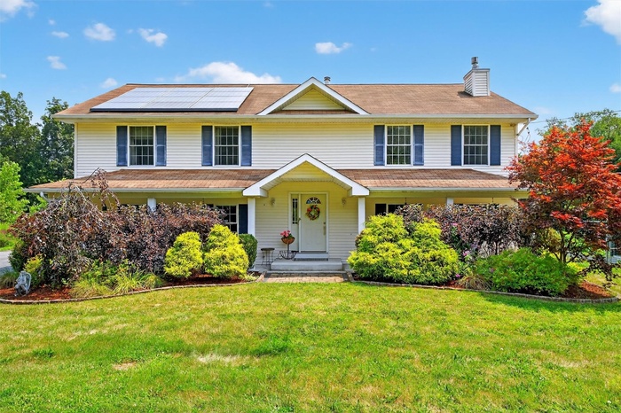 View of front facade featuring roof mounted solar panels, a chimney, a front lawn, and a porch