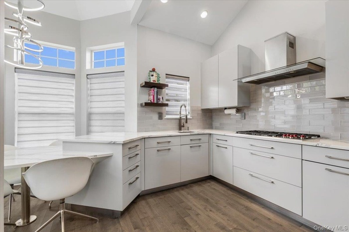 Kitchen featuring decorative backsplash, a sink, high vaulted ceiling, wall chimney exhaust hood, and dark wood-type flooring