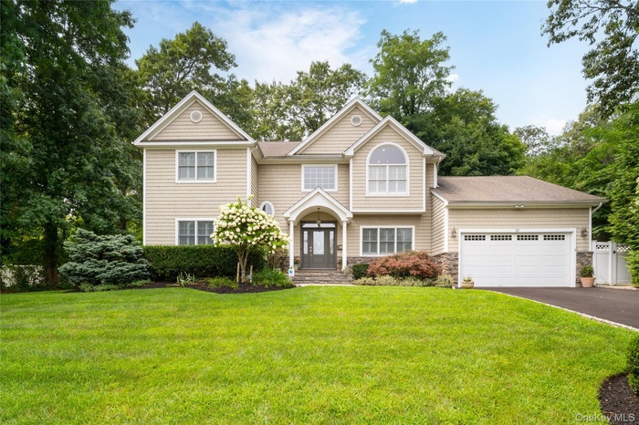 View of front facade with driveway, stone siding, a front yard, an attached garage, and view of wooded area