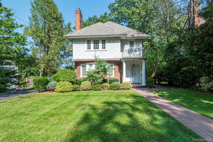 View of front of property with a balcony, a front yard, brick siding, a chimney, and a high end roof