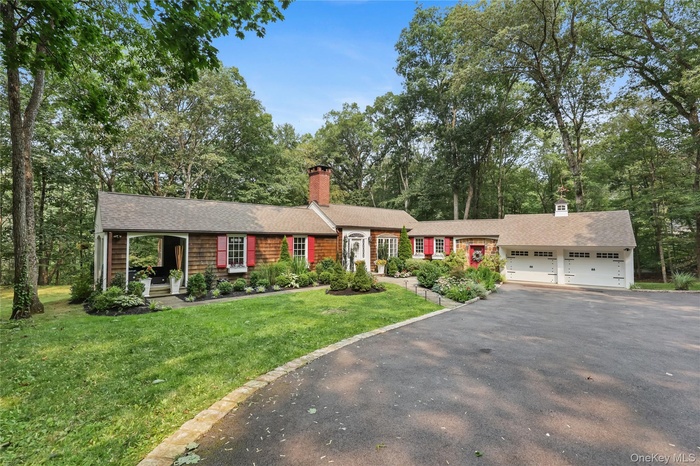 Single story home with a chimney, driveway, an attached garage, a front lawn, and view of wooded area