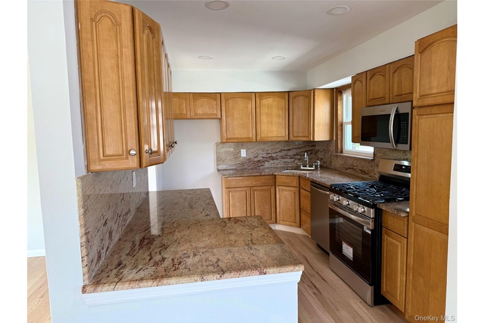 Kitchen with appliances with stainless steel finishes, light stone countertops, light wood finished floors, tasteful backsplash, and brown cabinets