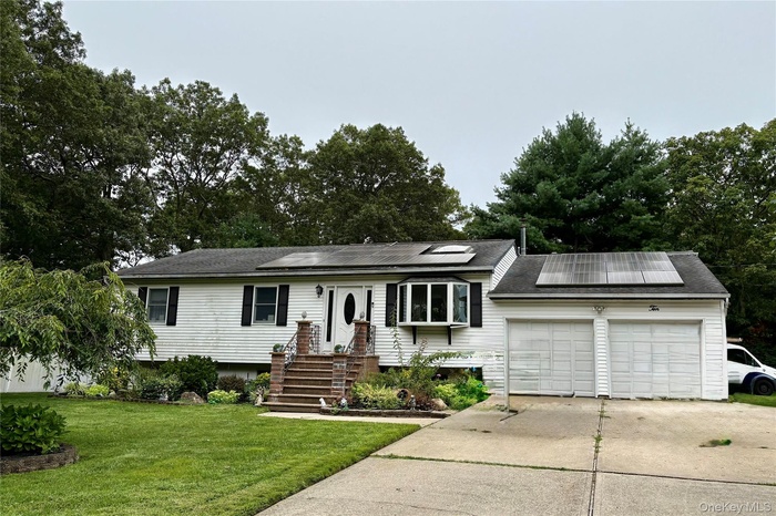 View of front facade with roof mounted solar panels, driveway, a front lawn, a garage, and view of scattered trees