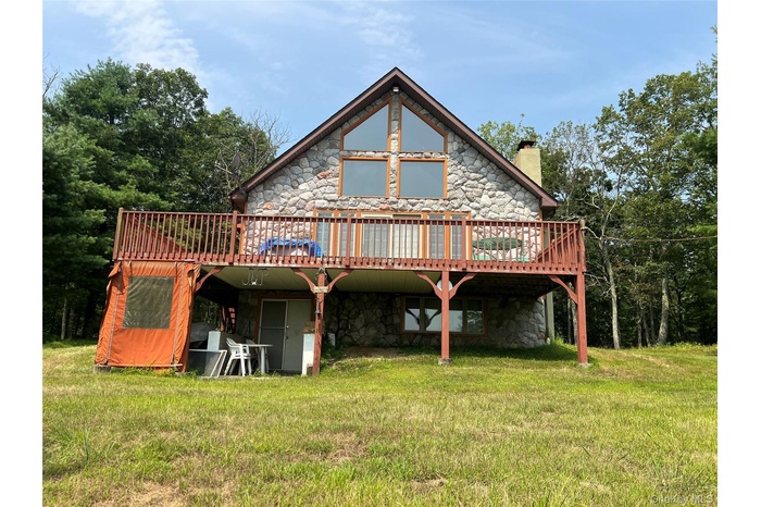 Back of property featuring a wooden deck, stone siding, a chimney, and a lawn