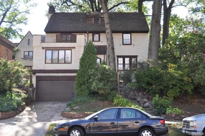 Tudor-style house with stucco siding, a garage, a balcony, a shingled roof, and driveway