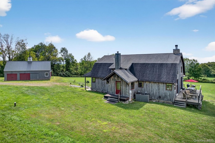 Front view of property featuring dirt driveway, a chimney, a lawn, a deck, and a shingled roof