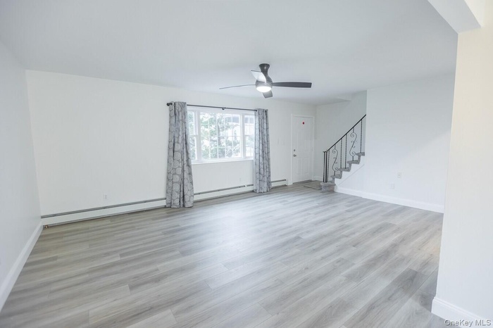 Unfurnished living room featuring stairway, light wood-style floors, a baseboard radiator, and ceiling fan