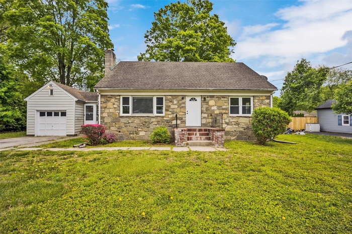 Cape Cod house featuring stone siding, a chimney, a shingled roof, a garage, and asphalt driveway