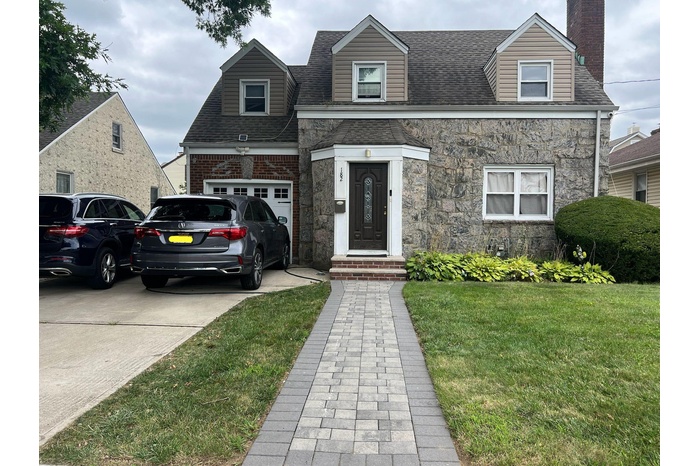 View of front of house featuring a chimney, concrete driveway, a front lawn, roof with shingles, and stone siding