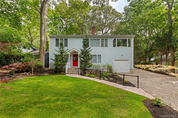 Colonial home featuring a garage, gravel driveway, view of wooded area, a chimney, and a front lawn