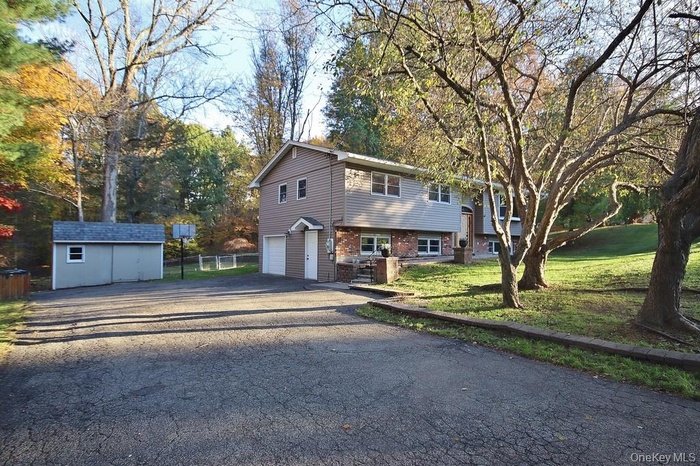 Raised ranch with asphalt driveway, brick siding, an attached garage, view of scattered trees, and a storage shed