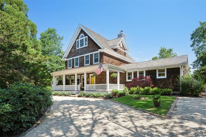 Shingle-style home with a porch, a chimney, gravel driveway, and a shingled roof