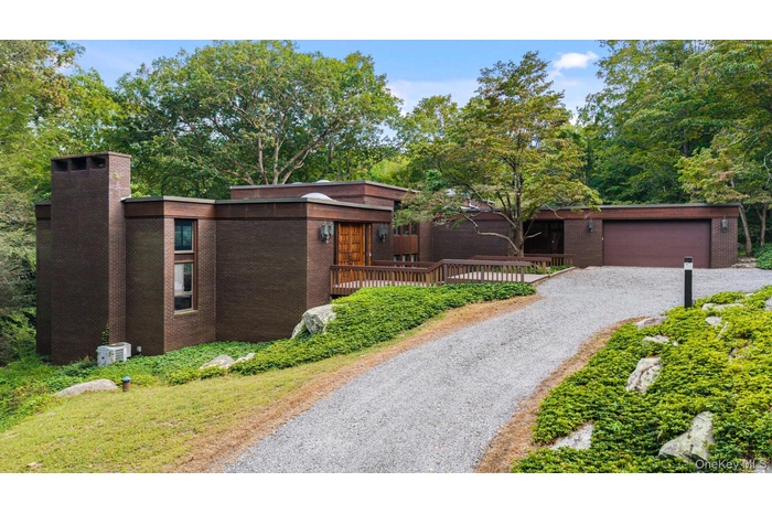 View of front of property featuring brick siding, gravel driveway, a chimney, a wooden deck, and an attached garage