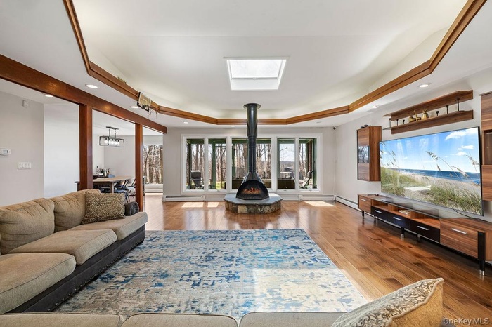 Living room with a raised ceiling, a wood stove, wood finished floors, a skylight, and a baseboard heating unit