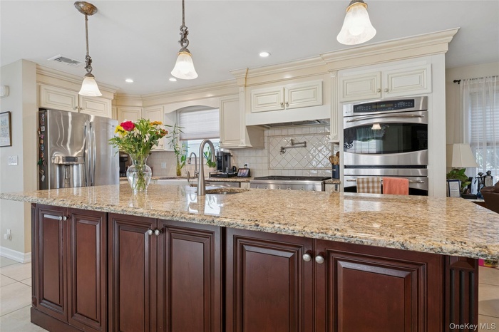 Kitchen featuring light tile patterned floors, appliances with stainless steel finishes, backsplash, hanging light fixtures, and light stone counterto