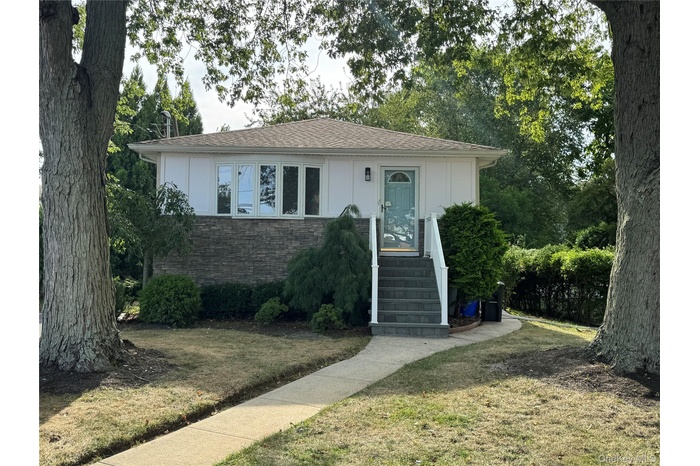 View of front of property featuring a front yard, stone siding, a shingled roof, and view of scattered trees