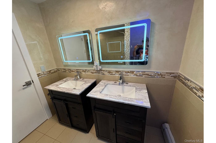 Full bath with tile walls, two vanities, a baseboard heating unit, light tile patterned floors, and a wainscoted wall