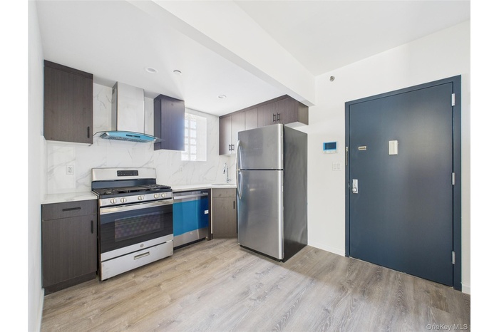 Kitchen featuring appliances with stainless steel finishes, wall chimney exhaust hood, decorative backsplash, light wood-style flooring, and dark brow