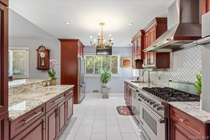 Kitchen with wall chimney range hood, high quality appliances, a chandelier, light tile patterned flooring, and tasteful backsplash