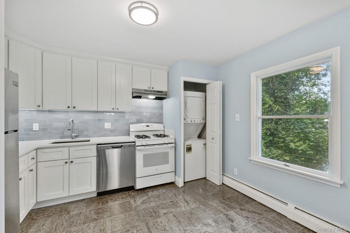 Kitchen featuring a baseboard heating unit, appliances with stainless steel finishes, backsplash, white cabinets, and stacked washer and clothes dryer