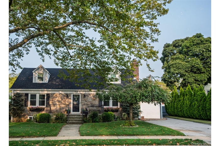 New england style home with driveway, a front lawn, a chimney, stone siding, and roof with shingles