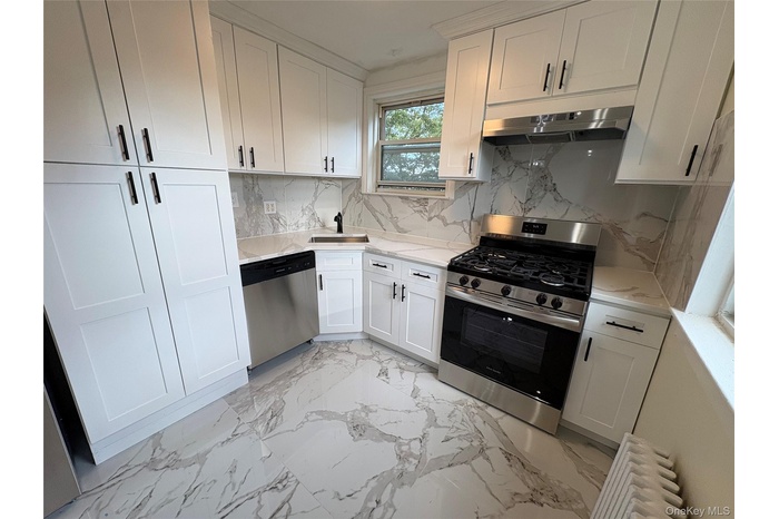 Kitchen with appliances with stainless steel finishes, decorative backsplash, radiator heating unit, under cabinet range hood, and light marble finish