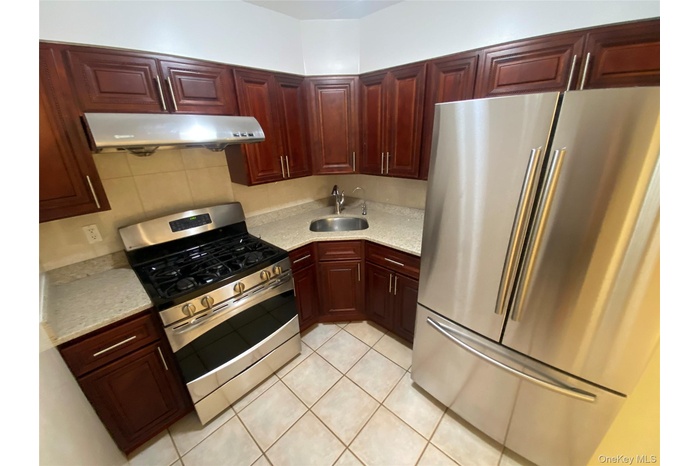 Kitchen with appliances with stainless steel finishes, light tile patterned flooring, under cabinet range hood, reddish brown cabinets, and light ston