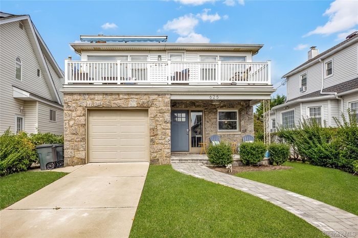 View of property featuring a front lawn, driveway, stone siding, a balcony, and an attached garage