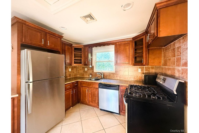 Kitchen with stainless steel appliances, glass insert cabinets, tasteful backsplash, brown cabinetry, and light tile patterned flooring
