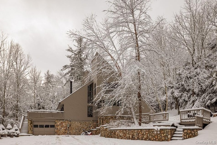 Snow covered property featuring stairway, a wooden deck, stone siding, and a garage