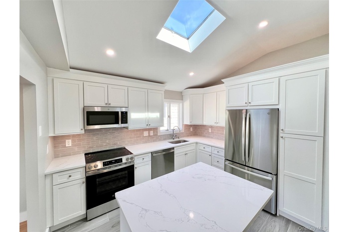Kitchen featuring vaulted ceiling, stainless steel appliances, light stone countertops, white cabinets, and decorative backsplash