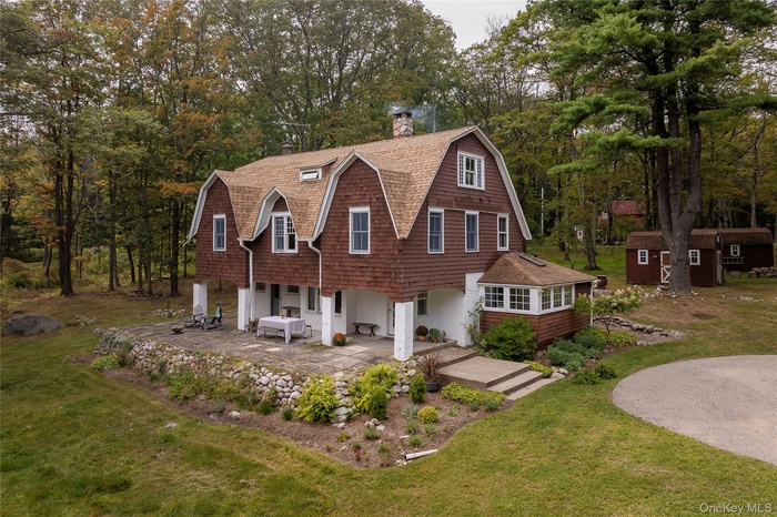 View of front facade with a gambrel roof, a patio, a chimney, and a shingled roof