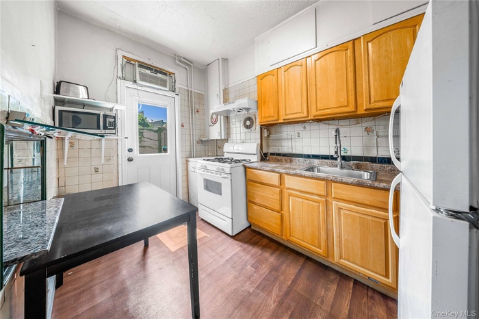Kitchen with white appliances, dark wood-style flooring, tasteful backsplash, dark stone counters, and under cabinet range hood