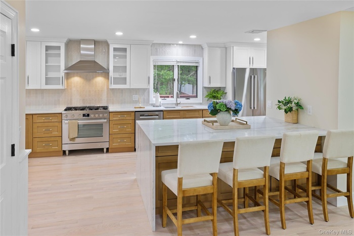Kitchen with a breakfast bar area, stainless steel appliances, recessed lighting, wall chimney exhaust hood, and light stone counters