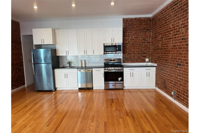 Kitchen with brick wall, white cabinets, appliances with stainless steel finishes, crown molding, and light wood-style flooring