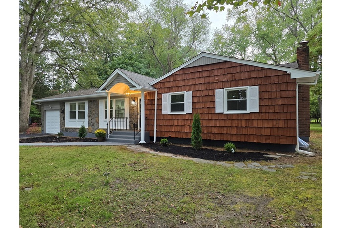 View of front facade with a chimney, a front lawn, an attached garage, view of wooded area, and driveway