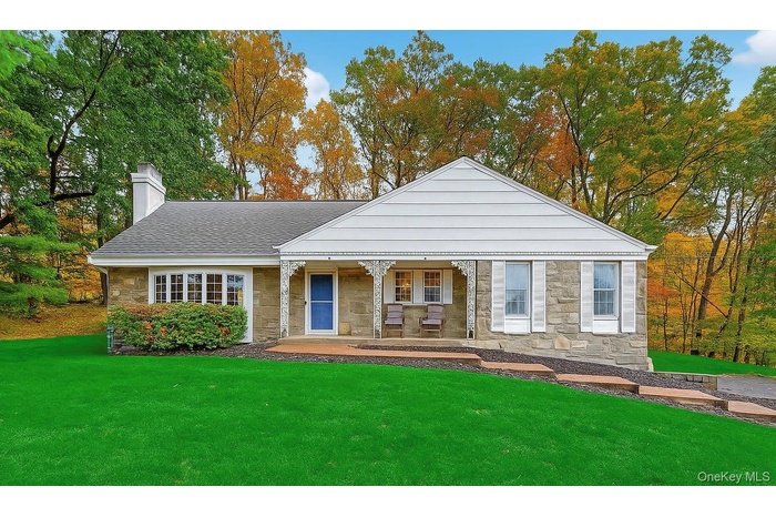 View of front facade with stone siding, a chimney, covered porch, a front lawn, and roof with shingles